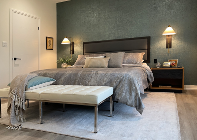Primary bedroom with custom headboard upholstered in suede. Brass Franklin side lights mounted on teal cork wallpaper. Bed and white leather bench on an area rug. Quartz topped end tables.